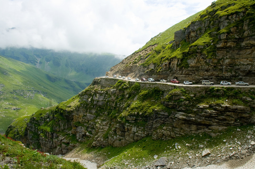 This is the road to the Rohtang Pass in northern India. This picture shows cars, mostly tourists visiting the area, queuing up on this narrow road.The photo emphasizes the paradoxon between pure nature in a remote mountain region and the heavy tourist traffic in that area.
